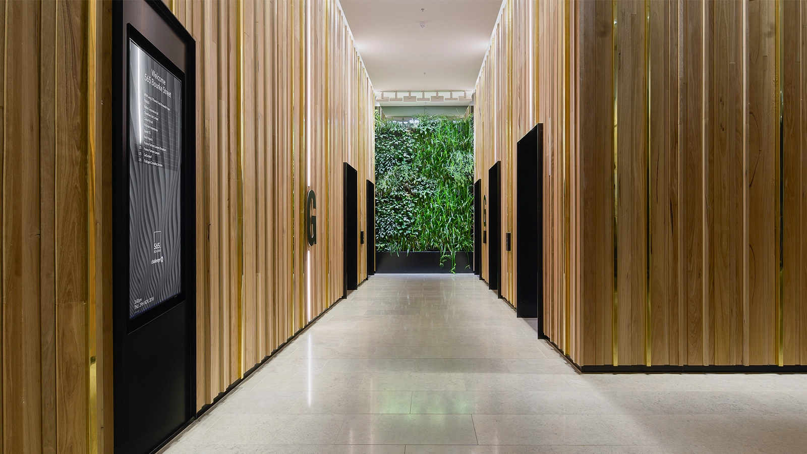 A wide angle shot of a wood panelled lift lobby with a recessed Wall Mounted Digital Directory to the left. A green wall of foliage can been seen at the rear of the lift lobby.