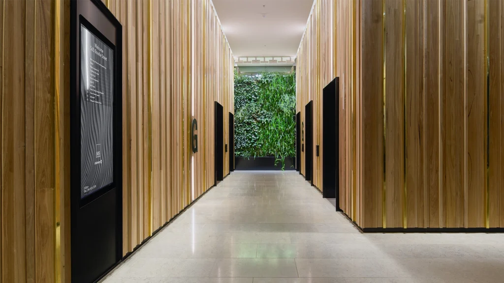 A wide angle shot of a wood panelled lift lobby with a recessed Wall Mounted Digital Directory to the left. A green wall of foliage can been seen at the rear of the lift lobby.