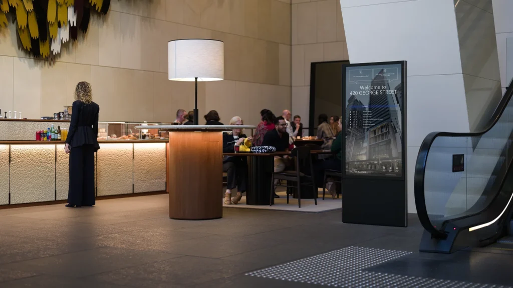 Freestanding Digital Tenant Directory in a crowded foyer of 420 George Street. People can be seens enjoying conversation at cafe.